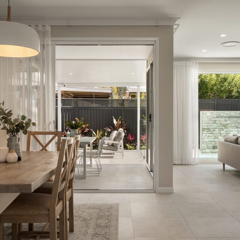 Stylish open-plan dining and living space with seamless indoor-outdoor flow, featuring a rustic wooden table, natural lighting, and a poolside patio. Designed by Jensen Constructions in Brisbane.